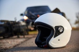 A white motorcycle helmet lies on the road with a blurred motorcycle and vehicle in the background.