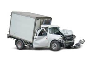 White delivery truck with significant front-end collision damage, including a crumpled hood and detached bumper. The cargo area appears unaffected. No background visible.