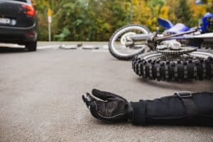 A motorcycle accident scene on a road, showing a person's gloved hand on the ground, with a fallen motorcycle and a car in the background.