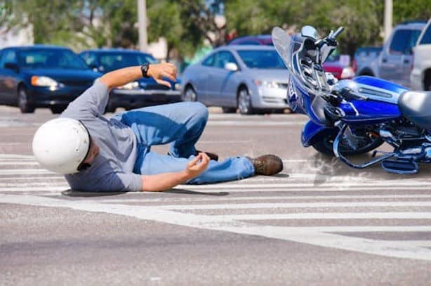 A motorcyclist wearing a helmet is falling on the road beside a fallen motorcycle at an intersection, with multiple cars in the background.