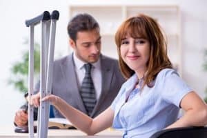Woman with crutches sits smiling at a desk, while a man in a suit reads a book in the background.