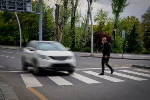 A white SUV quickly approaches a pedestrian in a crosswalk on a tree-lined street.
