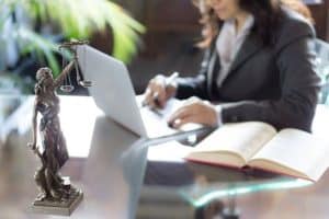 A person in a suit works on a laptop at a glass desk with an open book. A small statue of Lady Justice is in the foreground.