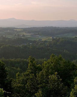 Rolling countryside landscape with dense green forests in the foreground, scattered houses in the middle, and hazy mountains under a soft pink sky in the background.