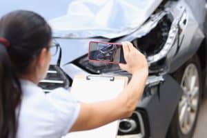 A woman photographs damage on a car with a smartphone while holding a clipboard. The car's front end is visibly dented.