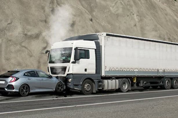 A car is crashed into the front of a large truck on a road with a rocky embankment beside it. Smoke is visible from the car's engine.