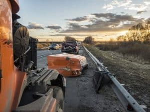 Roadside view of a crash involving a truck and a car. The car's rear is damaged, and debris is scattered around. Other vehicles are on the road, with a sunset sky in the background.