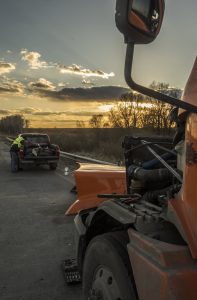 Truck with open hood and pickup truck on roadside at sunset; a person in a reflective vest checks the pickup's bed; grassy fields and trees in the background.
