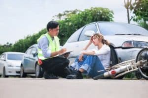 A man in a safety vest writes on a clipboard while kneeling beside a woman sitting on the ground next to a damaged bicycle and car.