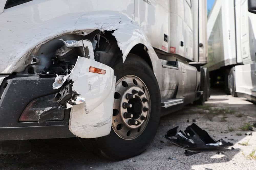 Close-up of a damaged white truck with a broken front bumper and headlight. Debris is scattered on the ground nearby.