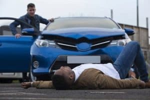 A blue car with front damage is stopped on the road. A man is lying in front of it, while another man stands by the car door, appearing concerned.