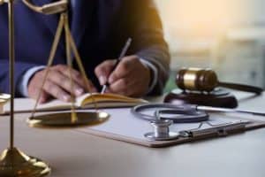 A person writing at a desk with a justice scale, stethoscope, clipboard, and gavel nearby.
