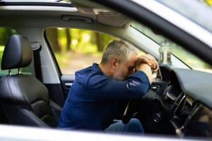 Man sitting in a car, leaning his head on the steering wheel with eyes closed, appearing tired or stressed.