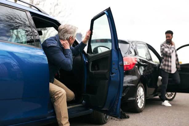 Two men assess a minor car accident. One man sits in a blue car, holding his neck, while the other stands near a black car. Both vehicles are on a road.