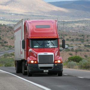 A red semi-truck drives on a highway through a desert landscape, with mountains visible in the distance.