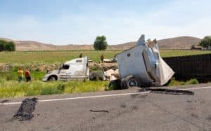 Overturned semi-truck and trailer off the side of a rural road, with debris scattered nearby. Workers in safety vests are inspecting the scene. Hills and trees are visible in the background.