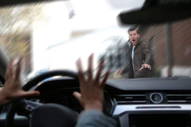 Person standing in front of a car with a surprised expression. Driver's hands visible on steering wheel, creating a scene of potential collision.