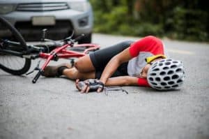 Cyclist in red and white jersey lying on the road beside a fallen red bike, wearing a helmet and gloves, with a car visible in the background.