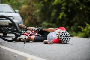 Cyclist wearing a helmet lies on the road near a fallen bicycle, with a car in the background.