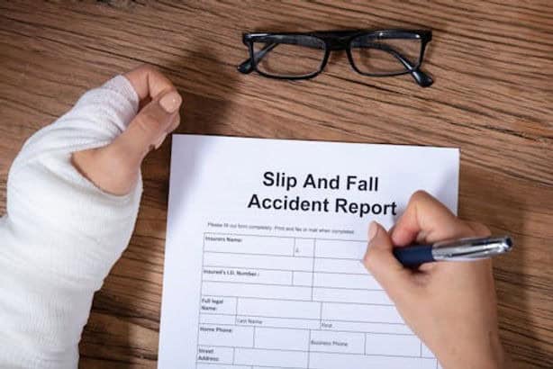 Individual with a bandaged hand fills out a "Slip and Fall Accident Report" form on a wooden table. Glasses are placed nearby.