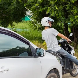 A person wearing a white helmet rides a scooter next to a white car, with trees and greenery in the background.