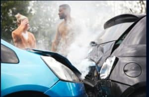 Two cars have collided, with smoke visible from the engines. Two individuals are standing nearby, one holding their head. Trees can be seen in the background.
