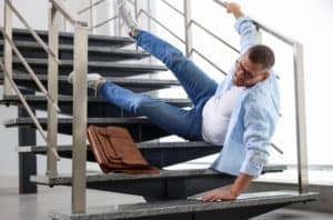 A man in jeans and a light blue shirt falls on a staircase while holding the railing. A briefcase lies nearby on the steps.