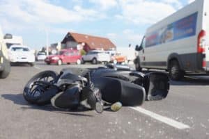 A fallen motorcycle lies on its side in the middle of a busy road with several vehicles and buildings in the background.