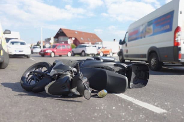 A fallen motorcycle lies on its side in the middle of a busy road with several vehicles and buildings in the background.