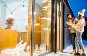 A man and woman in winter clothing stand outside a modern house with large windows. The man is opening a door while the woman looks on, holding mittens. A holiday wreath hangs on the door.