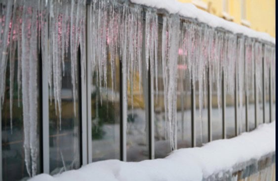 Icicles hang from a building's railing, with snow covering the surface. The background shows blurred reflections of windows and a warm glow.