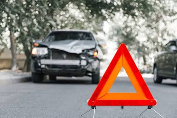 A car with front-end damage is parked on a road with a reflective warning triangle in the foreground. Trees and another vehicle are in the background.