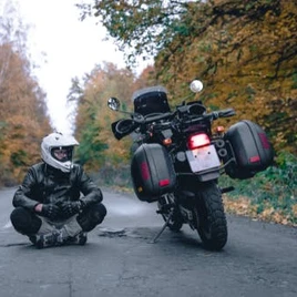 A person in a helmet and motorcycle gear sits beside a parked motorcycle on a forested road with autumn leaves.