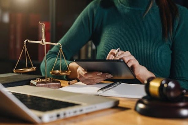 A person in a green sweater uses a tablet at a desk with a laptop, scale of justice, notebook, and gavel present.