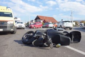 A fallen motorcycle lies in the middle of a road surrounded by several cars and an ambulance under a partly cloudy sky.
