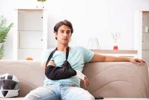 A person with a sling on their arm sits on a sofa in a bright room, next to a motorcycle helmet.