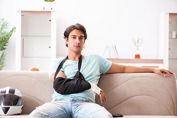 A person with a sling on their arm sits on a sofa in a bright room, next to a motorcycle helmet.