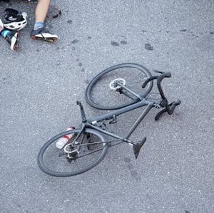 Bicycle on its side on an asphalt surface, with a person wearing cycling shoes and a helmet visible near the top left.