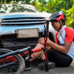 Cyclist sitting on the ground next to a fallen bike and car, holding his knee. He wears a helmet and cycling gear. Car is stationary with no visible license plate details.