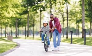 Adult helping a child ride a bicycle on a paved path in a park. The child is wearing a helmet and smiling, while the adult supports the bike from behind amidst green trees.