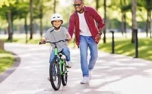 A smiling child rides a bike with training wheels on a paved path, wearing a helmet. An adult runs beside them, smiling, on a sunny day in a park with trees in the background.
