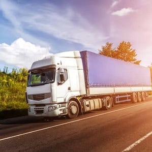 A white semi-truck with a blue trailer parked on a road under a sunny sky with trees in the background.
