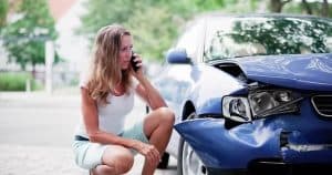 A woman crouches by a badly damaged blue car, talking on a cellphone, with a concerned expression.