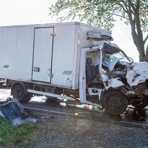 Damaged white truck with a crumpled front on a roadside, next to scattered debris, under a tree on a sunny day.