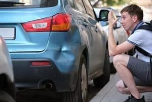 A person examines and takes a photo of a dent on the rear side of a parked blue car.