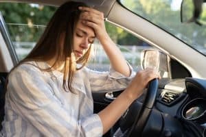 A woman sits in a car with her head resting on the steering wheel, looking distressed. Sunlight filters through the window.