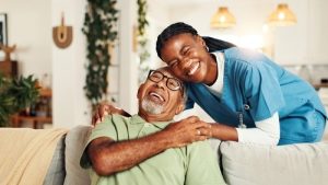 An elderly man sits on a couch smiling while a woman in scrubs, likely a caregiver or nurse, stands beside him, also smiling with her arm around his shoulder in a bright living room.