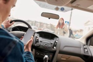 Driver inside a car is texting on a smartphone while a pedestrian, a woman, appears to be gesturing outside.
