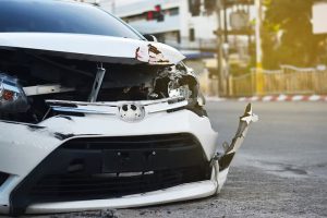 A white car with significant front-end damage is parked on a street near an intersection, with debris visible on the ground.