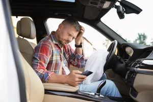 A man in a plaid shirt sits in a car, looking at his phone, with an airbag deployed in the passenger seat.